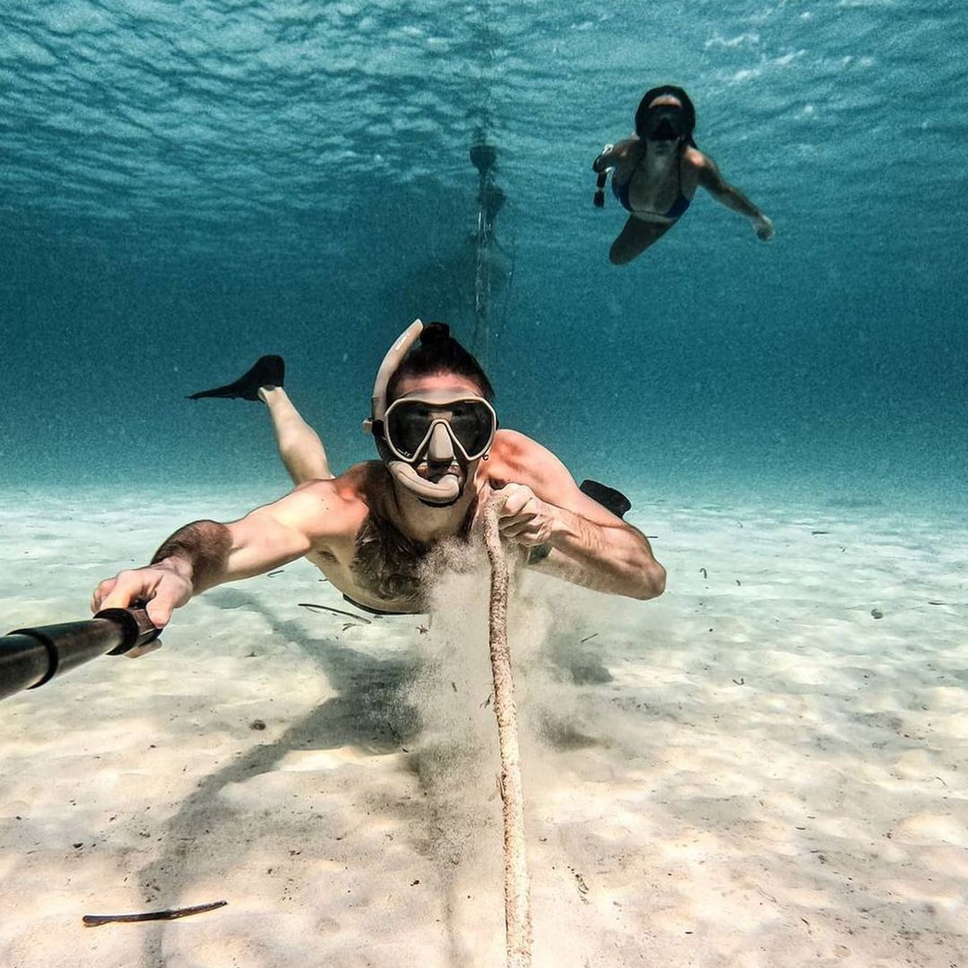 Un homme est sous l'eau en apnée. Il fait de la chasse sous-marine. Il porte un masque de plongée sous-marine Maxlux S Taupe et un tuba Spy. Une femme nage à la surface en arrière plan. L'eau est turquoise et le sable au fond est fin et doré. 